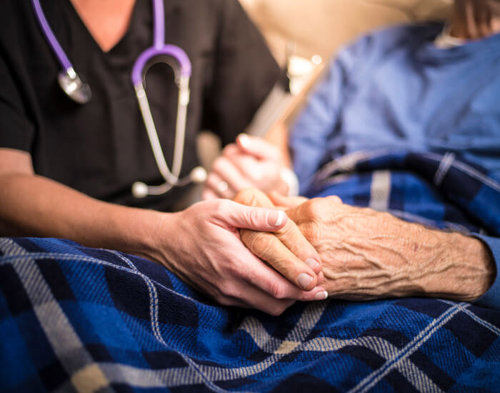 A hospice care worker holds the hand of a patient under a blanket in Torrance, CA.