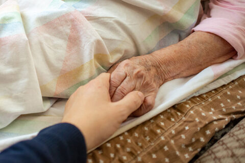 An elderly woman in hospice holds her daughter’s hand