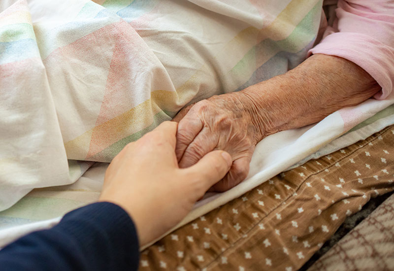 An elderly woman in hospice holds her daughter’s hand