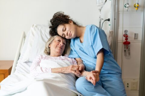 A nurse offering comfort and holding hands with a patient resting in a hospice bed in LA